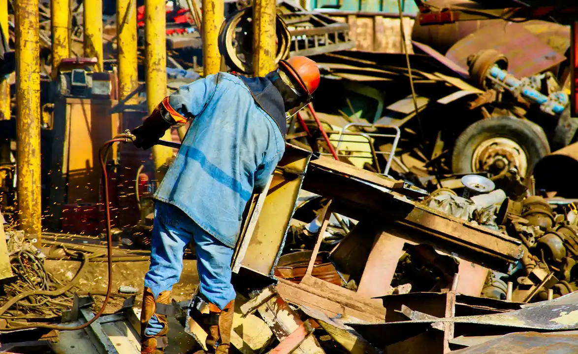 A worker in protective gear using a cutting torch on scrap metal amid piles of rusted machinery