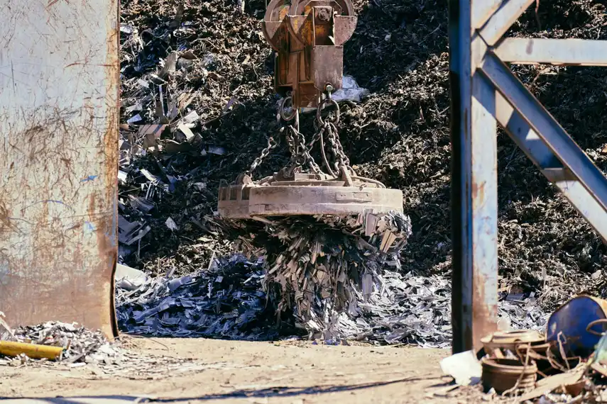 Aerial view of a large scrapyard with piles of crushed vehicles and a green grapple crane sorting scrap metal.