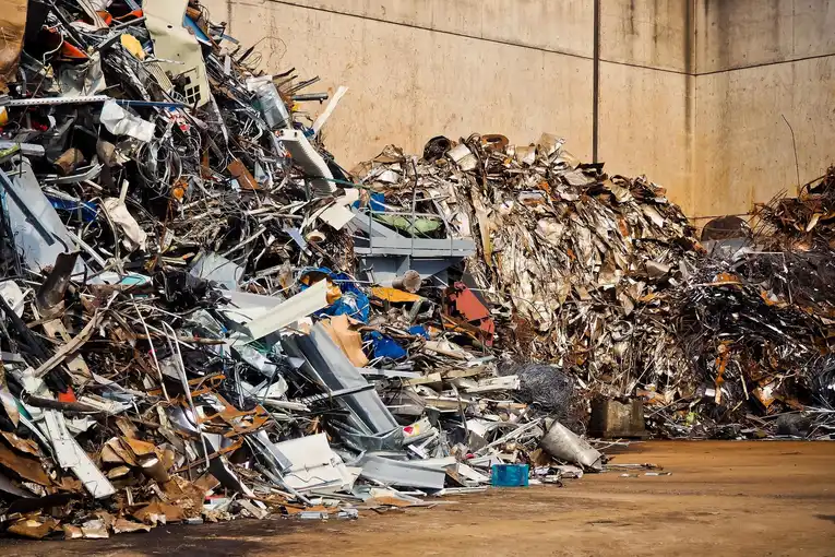 Large piles of mixed scrap metal, including twisted sheets and industrial debris, stacked in an outdoor recycling yard against a concrete wall.