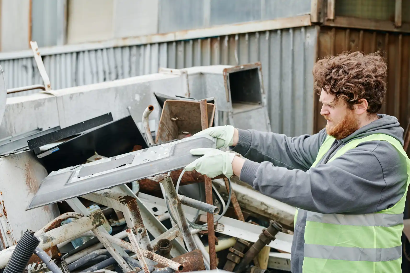 A worker in a hi-vis vest and gloves is sorting through scrap metal parts at an outdoor scrapyard.